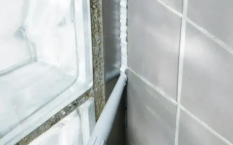 Applicator sprutar ny silikonfog längs en fog i badrummet, bredvid en duschvägg av glasblock.