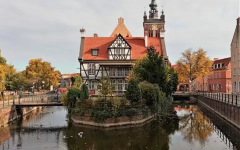 Urokliwy zakątek w **starym mieście Gdańsku**. Zabytkowa kamieniczka z czerwonym dachem i wieżą zegarową odbija się w spokojnej wodzie kanału.