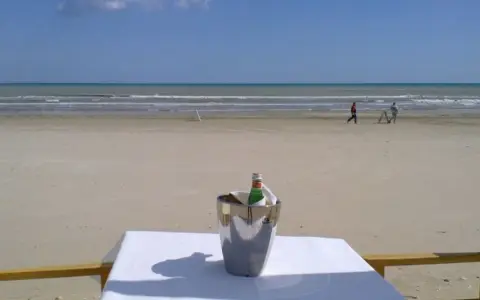 Coppia passeggia sulla spiaggia di Milano Marittima, con un secchiello del ghiaccio e una bottiglia su un tavolo in primo piano.