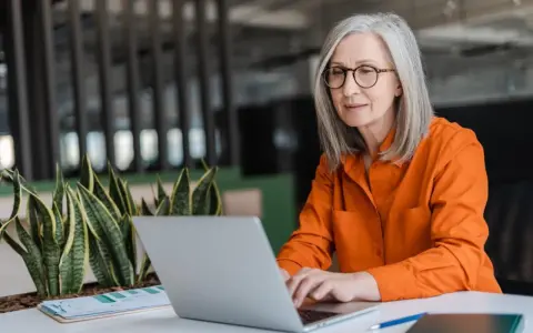 Mujer mayor con cabello plateado y gafas trabajando en su laptop, un ejemplo de jubilación activa.