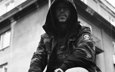Mężczyzna w kapturze i kurtce moro, z widocznym logo Timberland. Jego styl to ćpaj stajl czlonkowie, gotowy na miejskie wyzwania.
