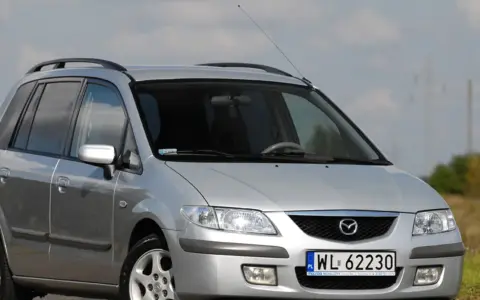 Srebrna Mazda Premacy z relingami dachowymi, zaparkowana na tle nieba i pól.