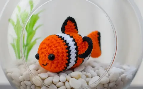 Adorable pez payaso de ganchillo en una pecera con piedras blancas y plantas verdes. ¡Perfecto para patrones de peces de ganchillo!
