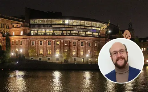 En man ler mot kameran framför Riksdagshuset i Stockholm, en symbol för representativ demokrati.