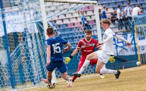 Piłkarz Michał Bartczak w białej koszulce próbuje strzelić gola, obrońca w niebieskiej koszulce z numerem 15 i bramkarz w czerwonej próbuje go zatrzymać.