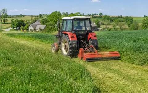 Czerwony traktor z mulczerem do c 330 kosi trawę na zielonym polu. W tle wiejskie domy i drzewa.