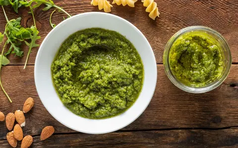 Pesto di rucola e mandorle in una ciotola bianca e un vasetto, con mandorle e rucola fresca intorno.