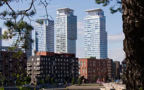 Höga, moderna skyskrapor reser sig bakom tegelbyggnader. En grönskande gren ramar in vyn, som om den undrar när sommaren startar i Sverige.