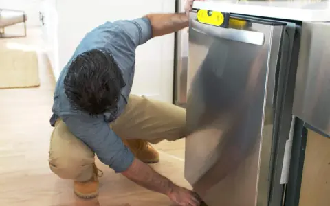 Man leveling a stainless steel dishwasher with a yellow bubble level, part of a guide on how to replace a dishwasher.