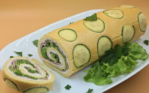 Rotolo di zucchine ripieno di prosciutto e insalata, decorato con fette di zucchina e prezzemolo su un piatto bianco.
