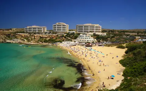En livlig strand med turkost vatten och gyllene sand vid Golden Bay, Malta. Flera hotell syns i bakgrunden.