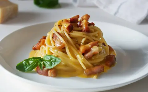 Pyszna carbonara z makaronem spaghetti, boczkiem i bazylią. W tle widać kawałek parmezanu, sugerując autentyczne carbonara skladniki.