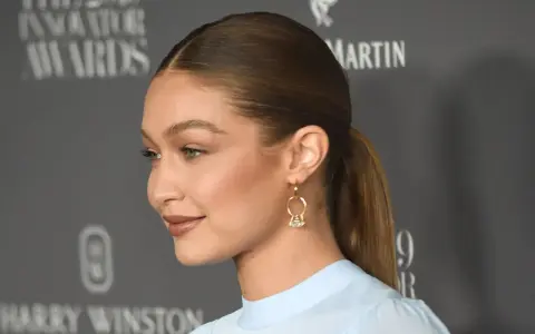 Gigi Hadid mit glatten, **haare nach hinten** gebundenen Haaren und einem dezenten Lächeln. Sie trägt ein hellblaues Oberteil und auffällige Ohrringe.