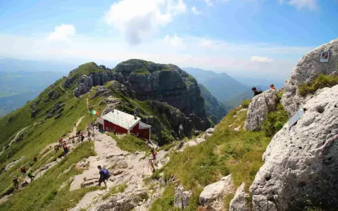 Sentiero panoramico verso il rifugio Lecco, con escursionisti su un crinale erboso e roccioso sotto un cielo azzurro.