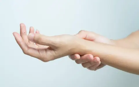 Dłoń z widocznym pieprzykiem na palcu, druga dłoń dotyka nadgarstka, gdzie wyczuwalna jest kość grochowata.
