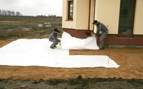 Dwóch robotników rozkłada białą geowłókninę, przygotowując tani taras na gruncie.