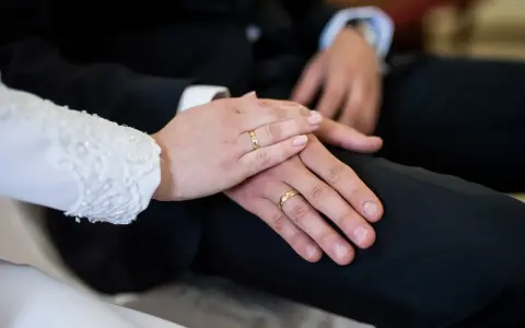 Dłonie nowożeńców z obrączkami na palcach, symbolizujące aklamację na ślub. Delikatny dotyk i złote kręgi mówią o miłości.