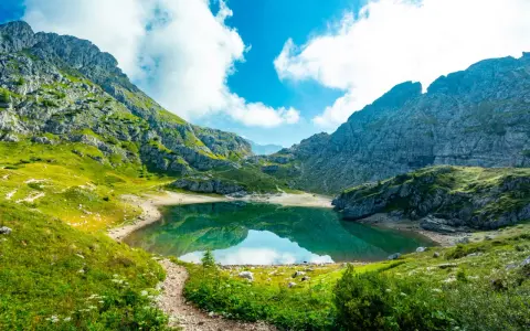 Sentiero panoramico tra le montagne, con un lago alpino dalle acque smeraldine che riflette il cielo azzurro e le nuvole bianche. Un perfetto percorso dolomiti.