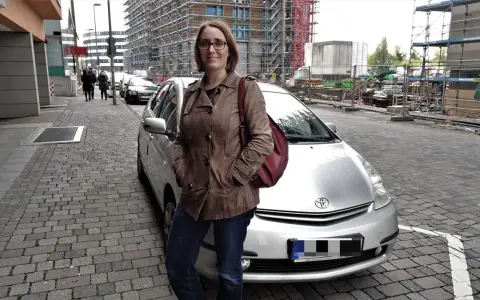 Frau mit brauner Lederjacke und roter Tasche steht neben silbernem Auto. Baustelle im Hintergrund. Sie ist free now fahrer.