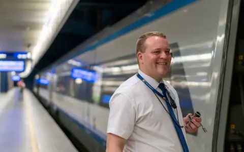 Uśmiechnięty konduktor PKP Intercity w białej koszuli i niebieskim krawacie stoi na peronie obok pociągu.