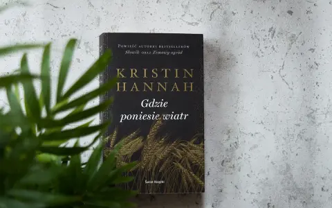 Okładka książki Kristin Hannah "Gdzie poniesie wiatr" na tle szarej ściany, z zielonymi liśćmi rośliny w zbliżeniu.