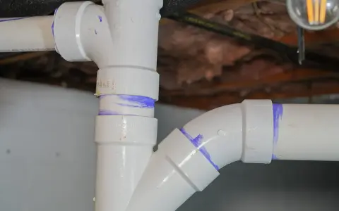 Close-up of various PVC pipe types, showing a vertical pipe connected to a horizontal pipe with a curved fitting, all marked with purple solvent cement.