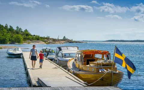 En kvinna och ett barn går på en brygga vid en sjö i Sverige. Båtar ligger förtöjda, en med svensk flagga. En perfekt familjesemester i Sverige.