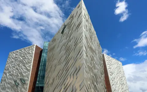 Nowoczesna bryła Muzeum Titanica w Belfaście, z geometrycznymi panelami i szklanymi elementami, pod błękitnym niebem.