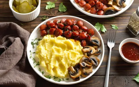 Talerz z purée ziemniaczanym, kiełbaskami i pieczarkami, obok miseczka z piklami i sosem.