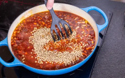 Eine Person rührt Linsen in eine köchelnde Bolognese-Sauce in einem blauen Topf.