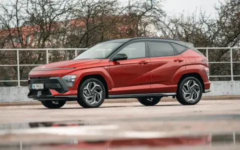 Czerwony Hyundai Kona Hybrid z czarnym dachem stoi na mokrym parkingu. Elegancki design i nowoczesne linie podkreślają jego hybrydowy charakter.