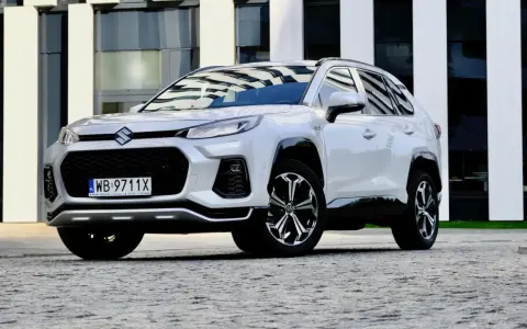 Biały Suzuki Across z czarnymi detalami stoi przed nowoczesnym budynkiem.