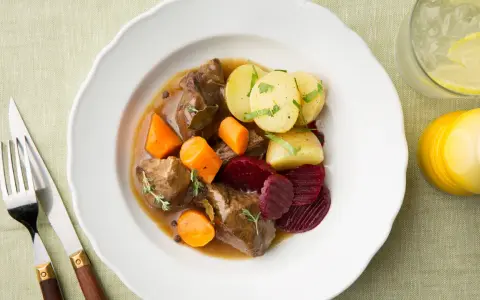 En tallrik med skånsk kalops, mört kött, morötter, potatis och rödbetor. Serveras med en drink och bestick.