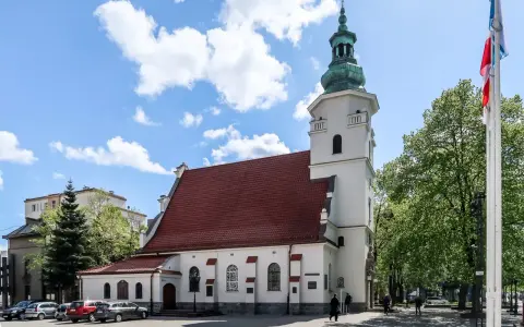 Biała bazylika pw. NMP Królowej Polski z czerwoną dachówką i zieloną wieżą, otoczona drzewami i budynkami.