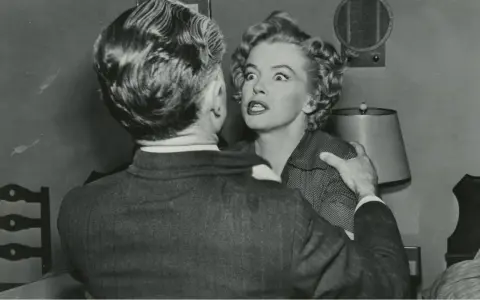 Marilyn Monroe med chockat uttryck i en scen från en av hennes filmer, hon håller fast vid en mans axel.