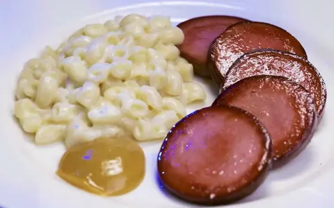 Stuvade makaroner snabbt vis med skivad falukorv och en klick senap på en vit tallrik.