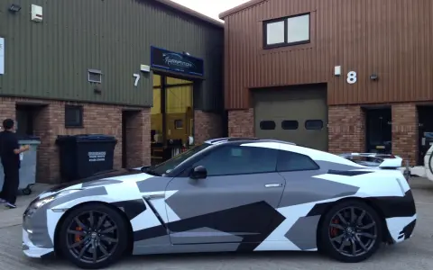 A Nissan GT-R with a striking geometric camouflage vinyl wrap for cars, parked outside a building with "WRAPTITION" signage.