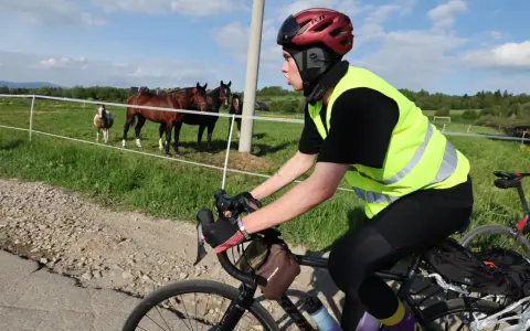 Kolarz w kamizelce odblaskowej i kasku, przygotowuje się do jazdy. Na łące pasą się konie. To jego rozgrzewka przed rowerem.