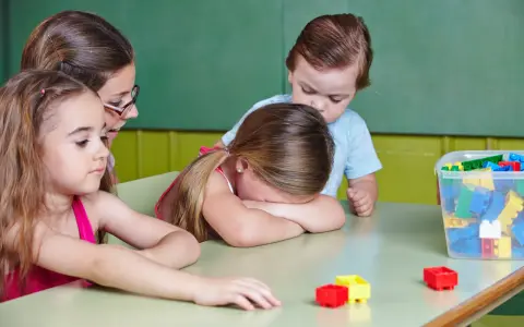 Smutna dziewczynka z płaczem ukrywa twarz na stole, podczas gdy inne dzieci i nauczycielka próbują ją pocieszyć. To obraz nieudanej adaptacji w przedszkolu.