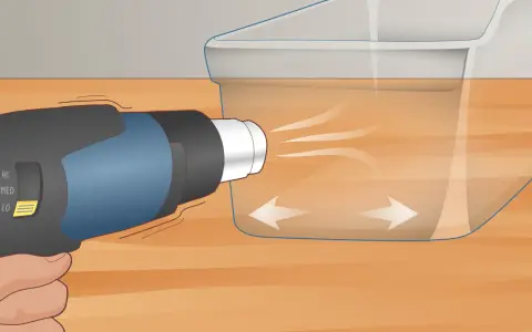 A heat gun is used on low setting to gently warm a scratched plexiglass surface, showing how to get scratches out of plexiglass.