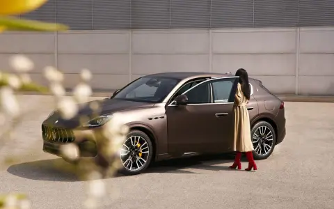 Elegancka kobieta w beżowej sukience i czerwonych kozakach zbliża się do brązowego Maserati SUV.