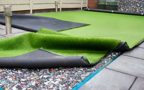 Instalación de césped artificial en terraza. Se ve el rollo de césped desplegado sobre una base de goma reciclada y baldosas.
