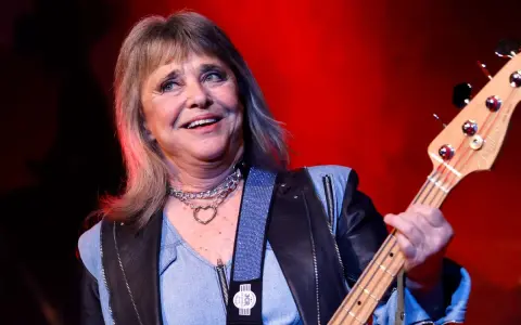 Suzi Quatro, die Rock-Ikone, spielt Bassgitarre. Sie trägt eine Lederjacke und eine Kette mit Herzanhänger. Ihr Alter ist nicht ersichtlich, aber ihre Energie ist spürbar.
