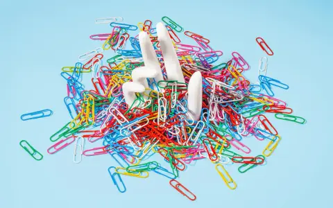 White hands emerge from a pile of colorful paperclips, symbolizing the struggle of how to stop being a workaholic and escape the daily grind.