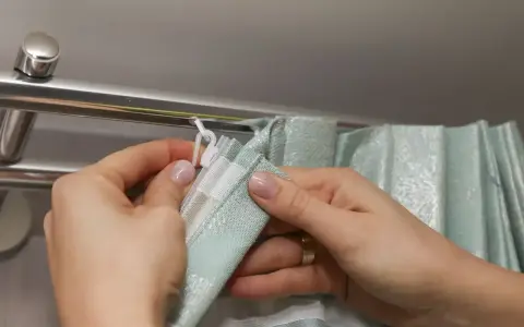 Dłonie wpinają białe haczyki flex do szyny karnisza, przygotowując zasłonę do zawieszenia.