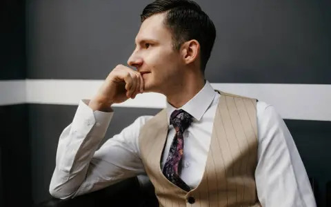 Elegancki mężczyzna w kamizelce, co zamiast marynarki, z dłonią na brodzie, wpatrzony w dal.