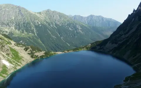 Górski krajobraz z jeziorem i stromymi zboczami. W oddali widać pasmo górskie, które przypomina najwyższy szczyt w Polsce.