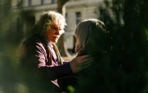 A man with white, curly hair and a mustache gently places his hand on another person's shoulder, offering a comforting response. This scene illustrates react vs respond examples.