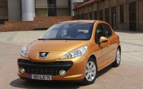Pomarańczowy Peugeot 207 1.4 benzyna zaparkowany na brukowanym placu przed ceglanym budynkiem.