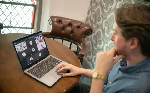Hombre en videollamada con sus equipos virtuales. Varios rostros aparecen en la pantalla de su portátil, conectados desde diferentes lugares.